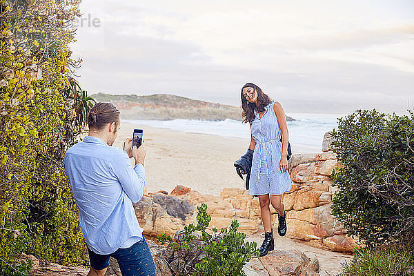 Young man with smart phone photographing girlfriend with ocean in background