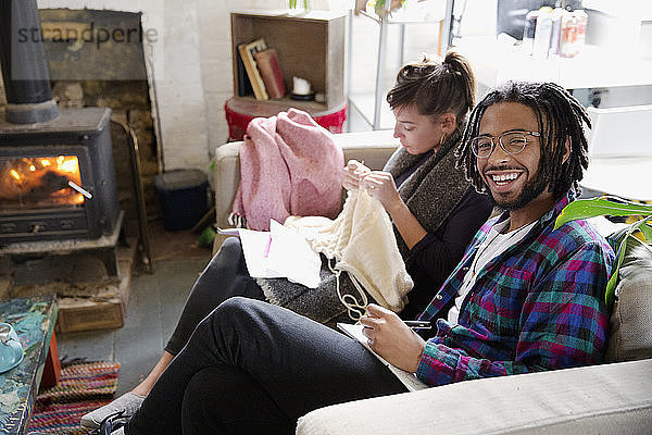 Porträt lächelnder junger Mann  der auf dem Sofa neben seiner strickenden Freundin in ein Notizbuch schreibt
