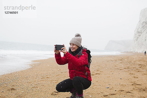 Frau in warmer Kleidung mit Fotohandy am verschneiten Strand