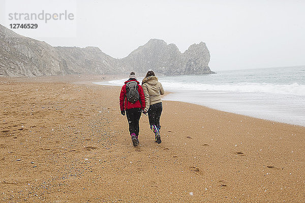Mutter und Tochter in warmer Kleidung spazieren am verschneiten Winterstrand am Meer