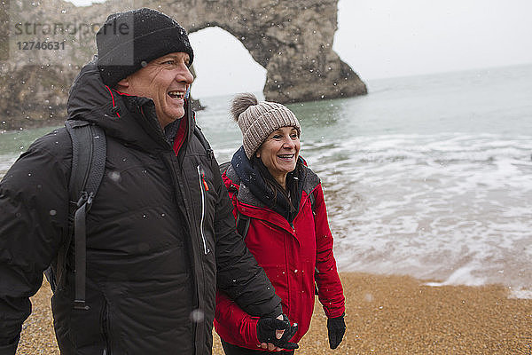 Glückliches Paar in warmer Kleidung spaziert am verschneiten Meeresstrand