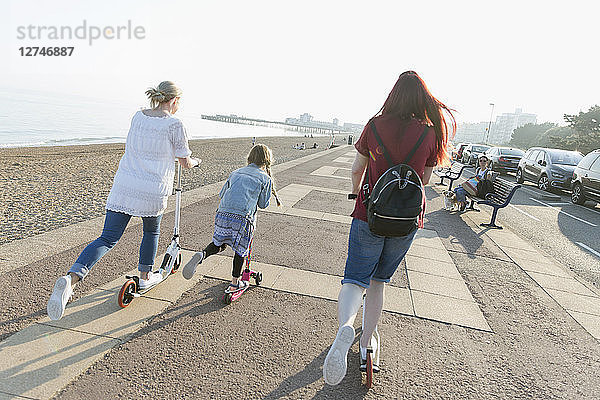 Lesbian couple with daughter riding push scoters on sunny beach boardwalk