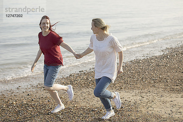 Playful lesbian couple holding hands and running on sunny beach
