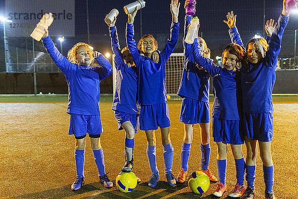 Porträt einer selbstbewussten Mädchenfußballmannschaft mit Wasserflaschen  die nachts auf dem Spielfeld jubeln