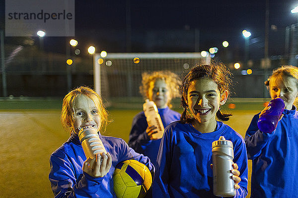 Porträt einer lächelnden Mädchenfußballmannschaft  die eine Trainingspause einlegt und nachts auf dem Spielfeld Wasser trinkt