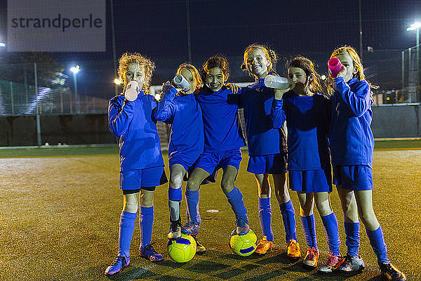 Porträt einer selbstbewussten Mädchenfußballmannschaft  die nachts auf dem Spielfeld Wasser trinkt