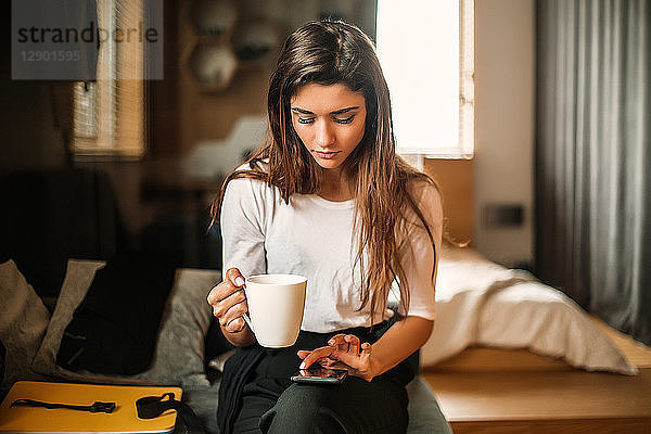 Frau mit Tasse benutzt Smartphone im Schlafzimmer