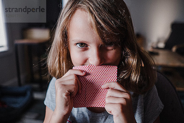 Mädchen versteckt sich hinter Spielkarten im Wohnzimmer  Porträt