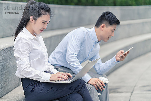 Junge Geschäftsfrau und junger Geschäftsmann mit Laptop und Blick auf Smartphone am Stadtsitz  Shanghai  China