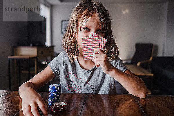 Mädchen  das sich hinter Spielkarten mit Spielchips am Tisch versteckt  Portrait