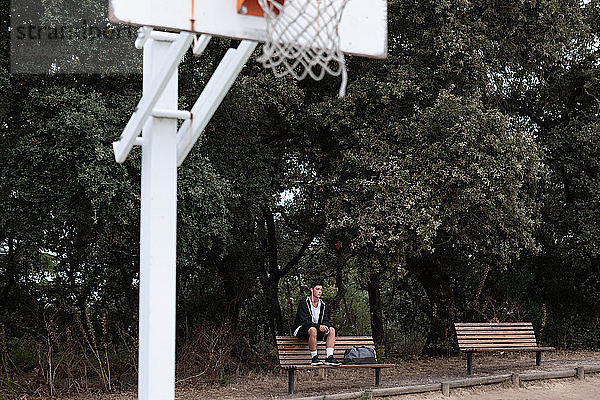 Männlicher jugendlicher Basketballspieler sitzt auf einer Parkbank am Basketballfeld