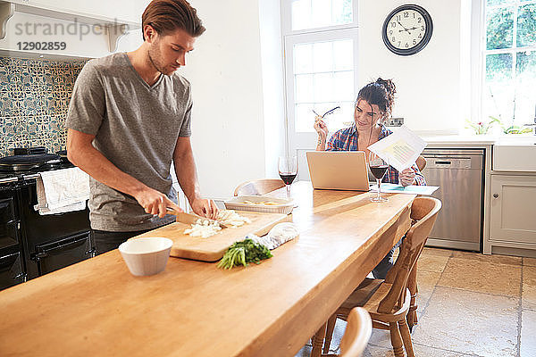 Mann bereitet Gemüse am Küchentisch zu  Freundin benutzt Laptop