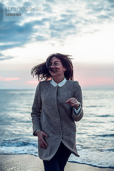 Porträt einer jungen Frau am Strand stehend  mit dem Rücken zum Ozean  der Wind weht ihr Haar  lächelnd