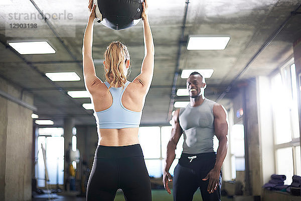 Trainer beobachtet weiblichen Klienten beim Heben eines Medizinballs im Fitnessstudio