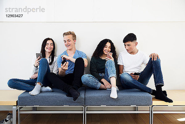 Vier Gymnasiasten auf Sitzgelegenheiten mit Blick auf Smartphones