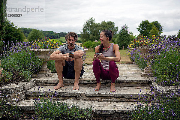 Mann und Frau üben Yoga im Garten und schauen sich auf der Terrasse Smartphones an