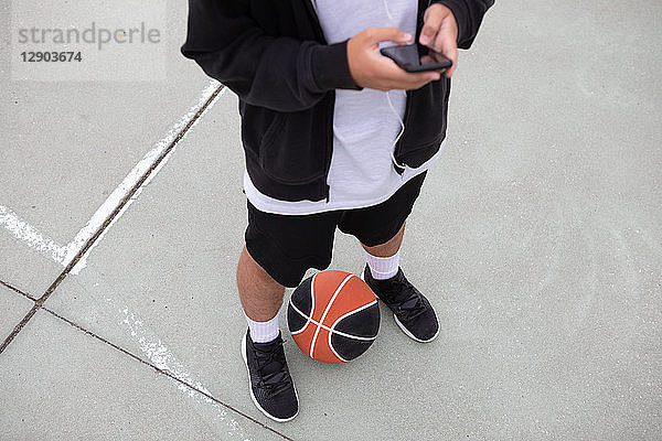 Männlicher jugendlicher Basketballspieler steht auf dem Basketballplatz und schaut auf ein Smartphone  Taille nach unten