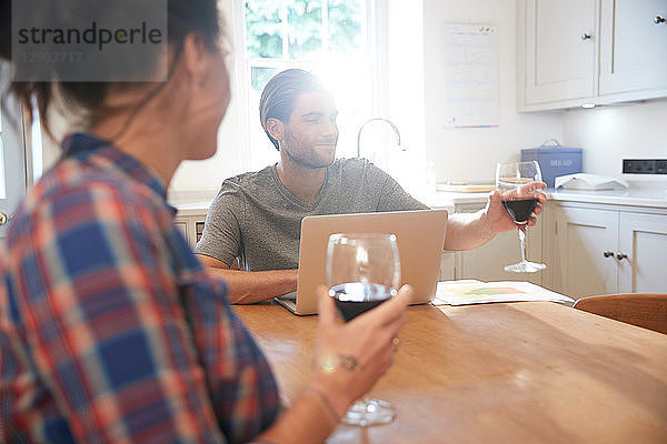 Ehepaar am Küchentisch trinkt Rotwein  während es einen Laptop benutzt