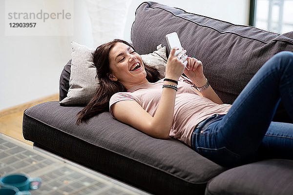 Teenager-Mädchen lacht auf Sofa und schaut auf Smartphone