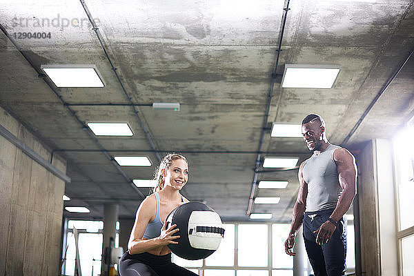 Trainer beobachtet weiblichen Klienten beim Heben eines Medizinballs im Fitnessstudio