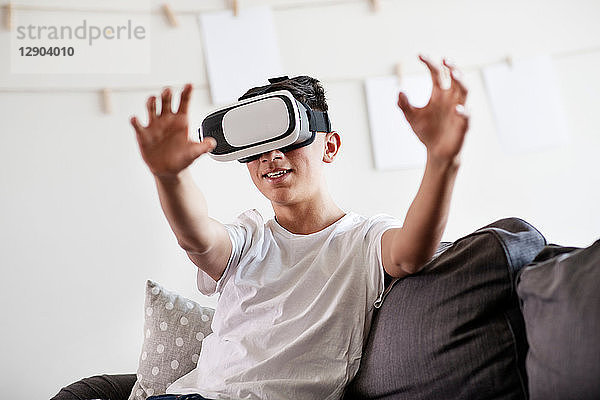 Teenager-Junge auf dem Sofa  der durch ein Virtual-Reality-Headset schaut und die Hand ausstreckt