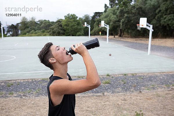 Männlicher jugendlicher Basketballspieler  der auf dem Basketballplatz aus einer Wasserflasche trinkt