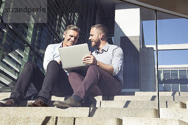 Two smiling businessmen sitting on stairs using laptop