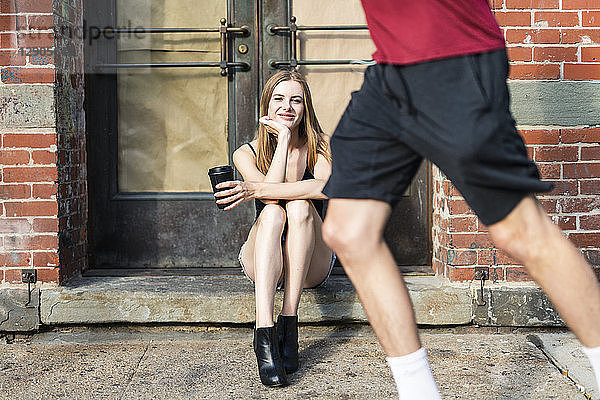 Young woman sitting in the street on a doorstep  holding a cup of coffee  jogger passing