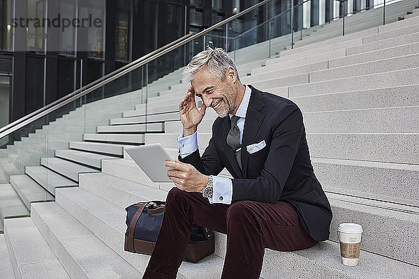 Fashionable businessman with travelling bag and coffee to go sitting on stairs using tablet