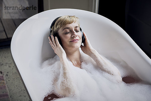 Blond woman taking bubble bath in a loft listenung music with headphones