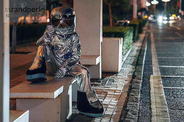 Spaceman sitting on bench at a bus stop at night holding cell phone