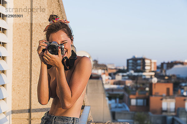 Happy topless young woman with camera taking pictures on balcony