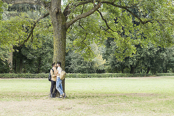 Kissing young gay couple leaning against tree in city park