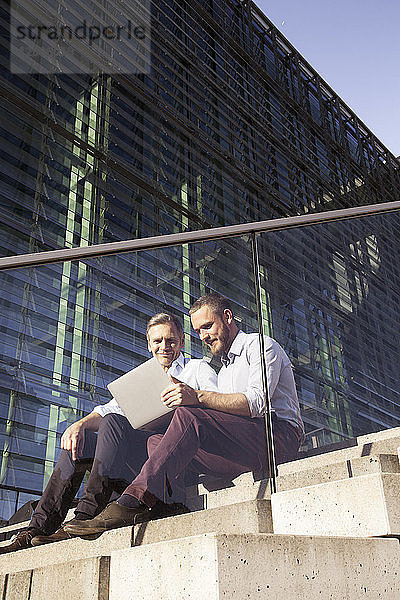 Two smiling businessmen sitting on stairs using laptop