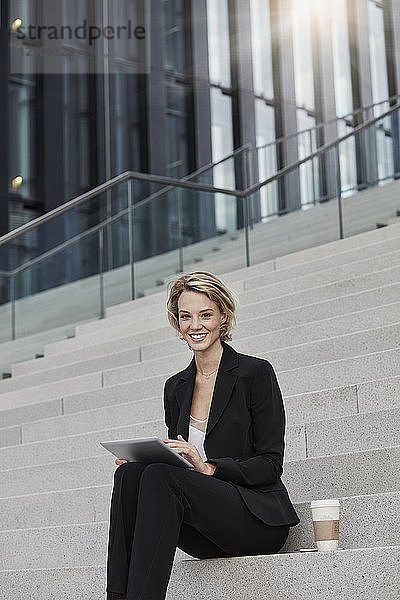 Portrait of smiling businesswoman with tablet and coffee to go sitting on stairs in front of modern office building