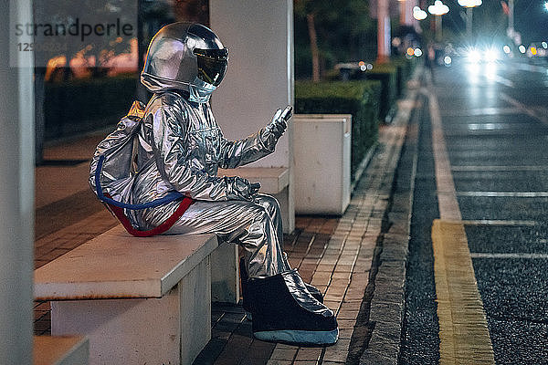 Spaceman sitting on bench at a bus stop at night holding cell phone