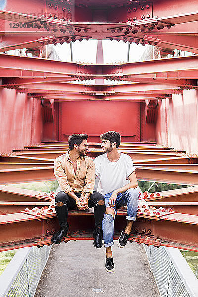 Young gay couple in love sitting on steel girders of a footbridge looking at each other