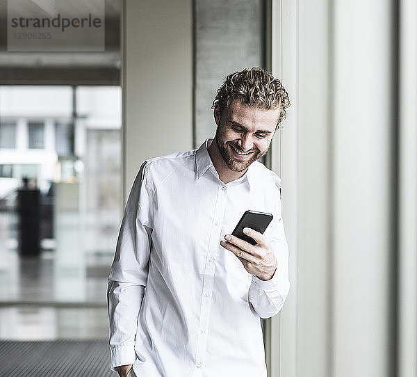 Smiling young businessman looking at cell phone at the window in office