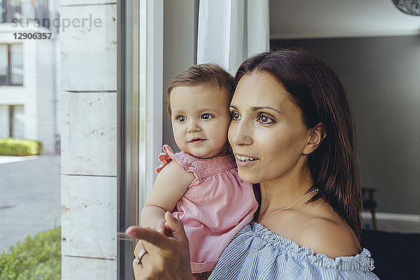 Smiling mother looking out of window with baby daughter at home