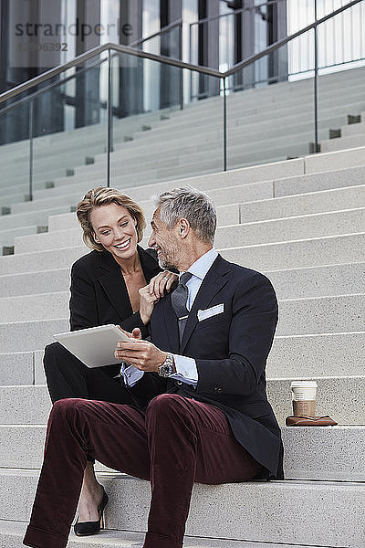 Two smiling business people with tablet sitting together on stairs