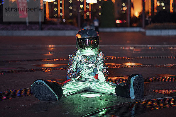 Spaceman sitting at a lamp on a city square at night warming his hands