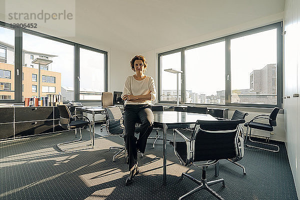 Successful businesswoman sitting on desk in her office with arms crossed