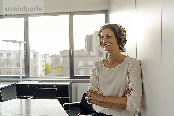Businesswoman leaning against wall in office  with arms crossed  smiling