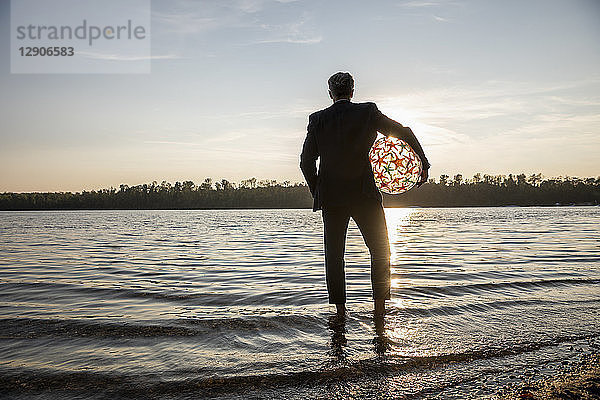 Back view of businessman with beach ball standing at lakeshore watching sunset