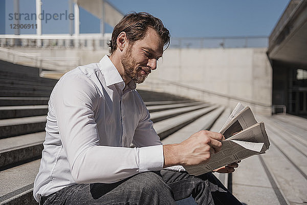Smiling businessman sitting on stairs reading newspaper