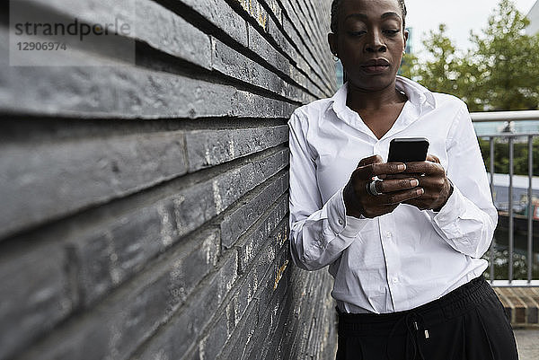 Serious businesswoman leaning against wall looking at smartphone