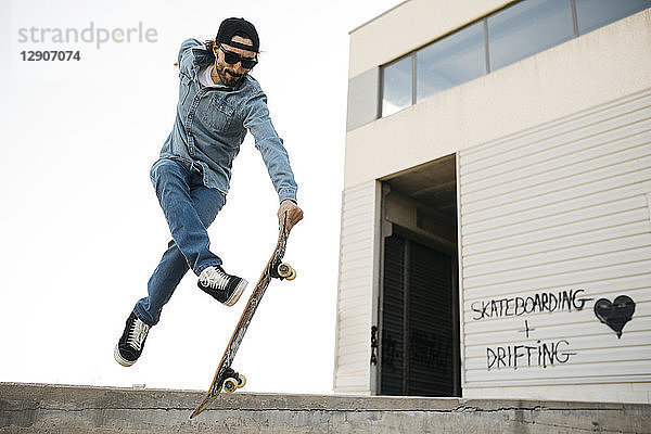 Trendy man in denim and cap skateboarding  doing jump with skateboard from concrete ramp