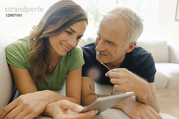 Smiling mature couple lying on couch at home sharing tablet
