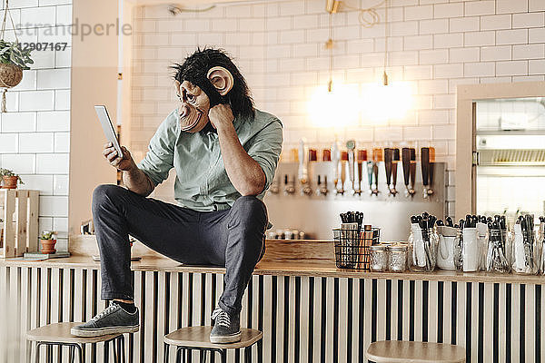 Man wearing monkey mask  sitting on counter of a bar  using digital tablet