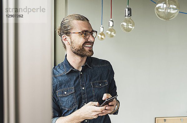 Smiling young businessman holding cell phone at the window in office